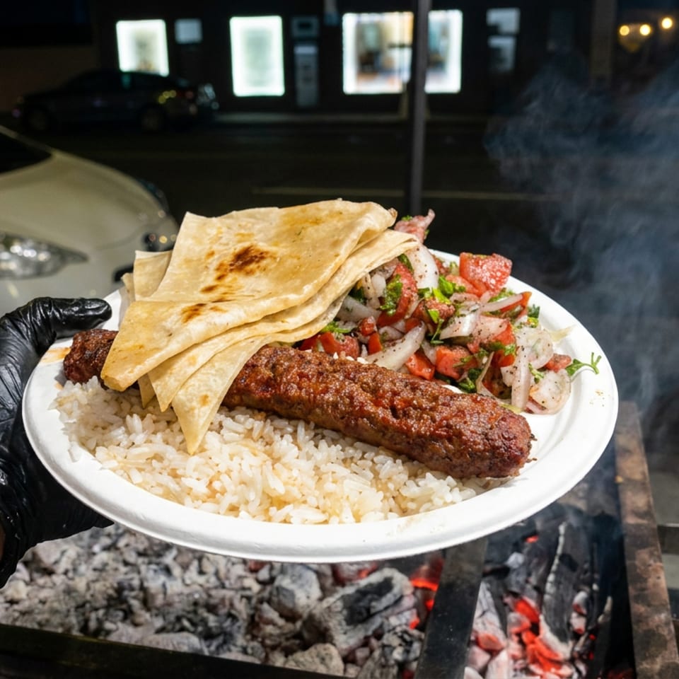 Plate of grilled kebab, rice, flatbread, and chopped salad held over a smoky charcoal grill at night.