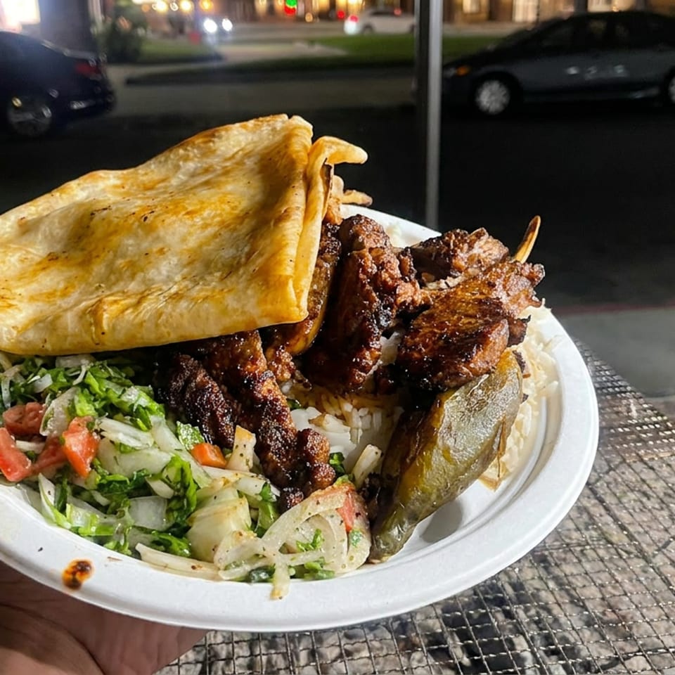 Plate of grilled meat over rice with salad, roasted vegetables, and flatbread, held outdoors beside a street.