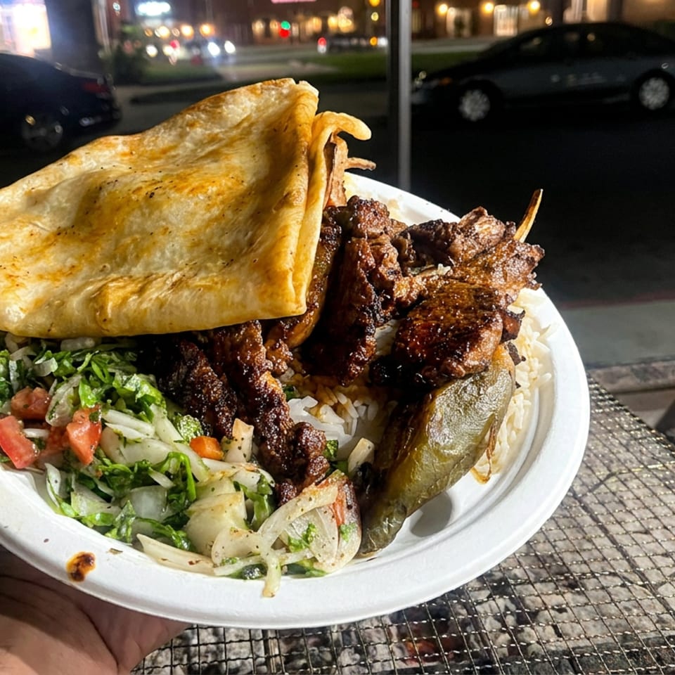 Plate of grilled meat over rice with salad, pickled vegetables, and flatbread, held outdoors beside a street grill.