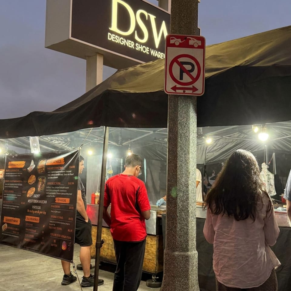 Customers queue at a lit outdoor food stall beneath a canopy at dusk, with a menu board and nearby Designer Shoe Warehouse sign.