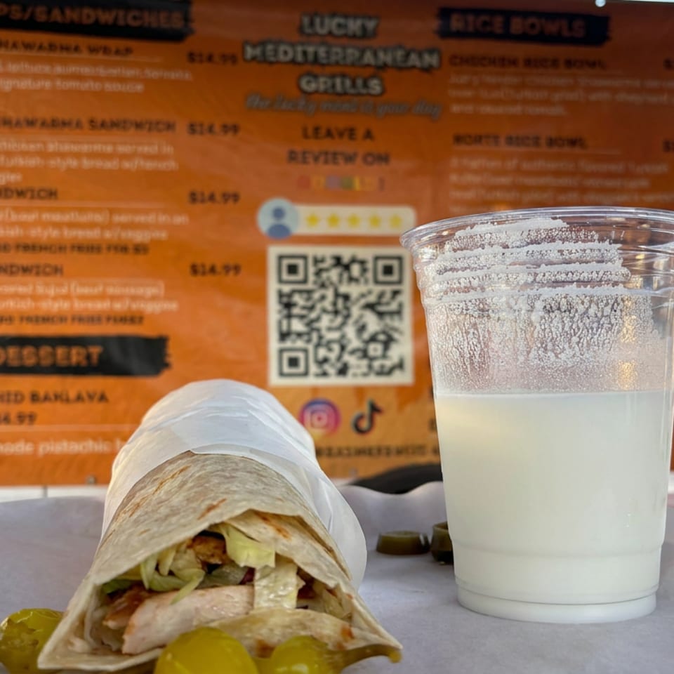 Chicken shawarma wrap and a cup of creamy white drink displayed before a Mediterranean grill menu.