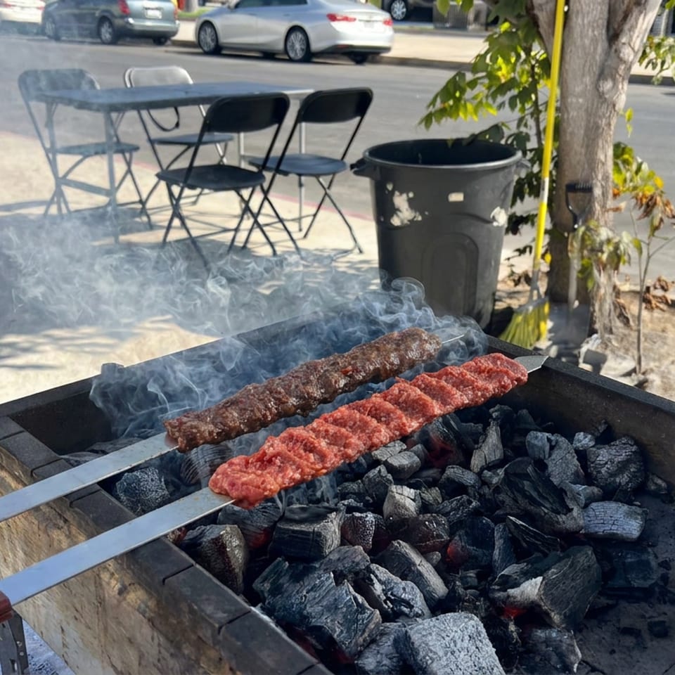 Two skewers of minced meat grilling over charcoal in a smoking outdoor barbecue beside a sidewalk.