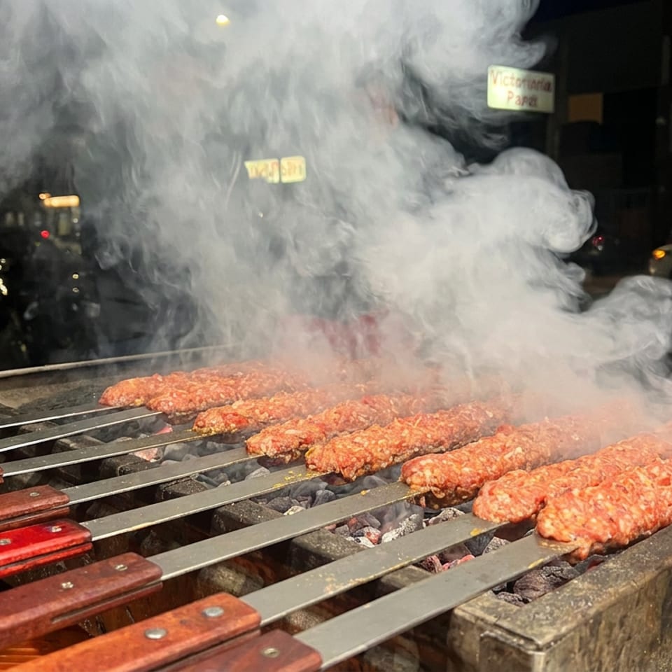 Smoky outdoor grill loaded with minced meat skewers, while a man in a skull shirt records nearby at night.